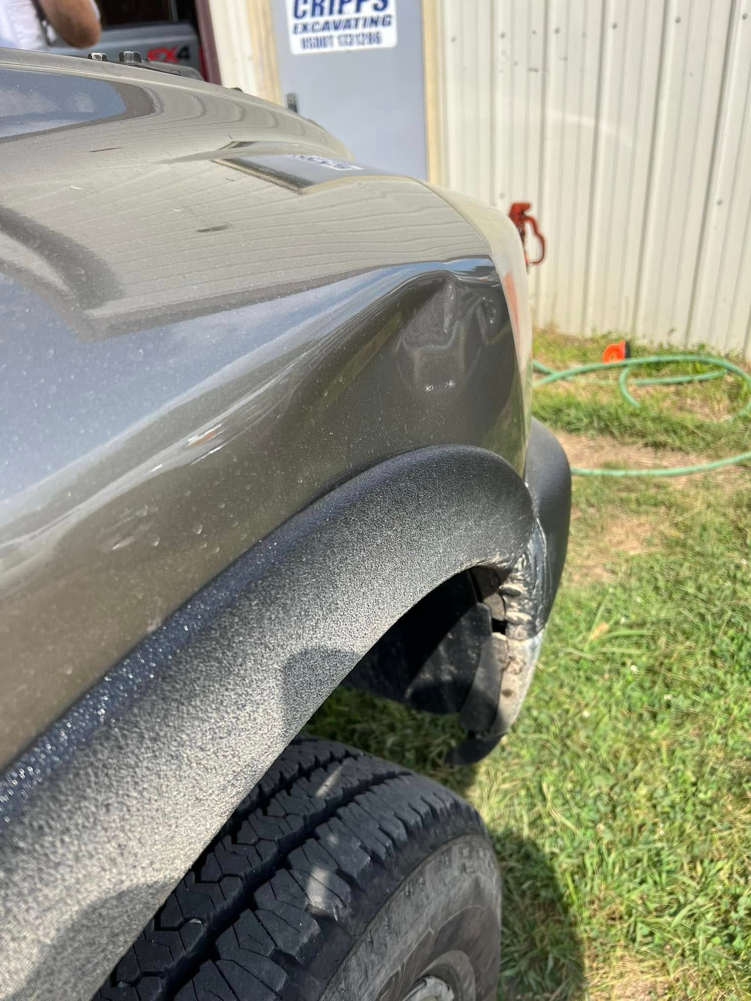 Close-up of a gray truck’s dented front fender and wheel near a fence outdoors