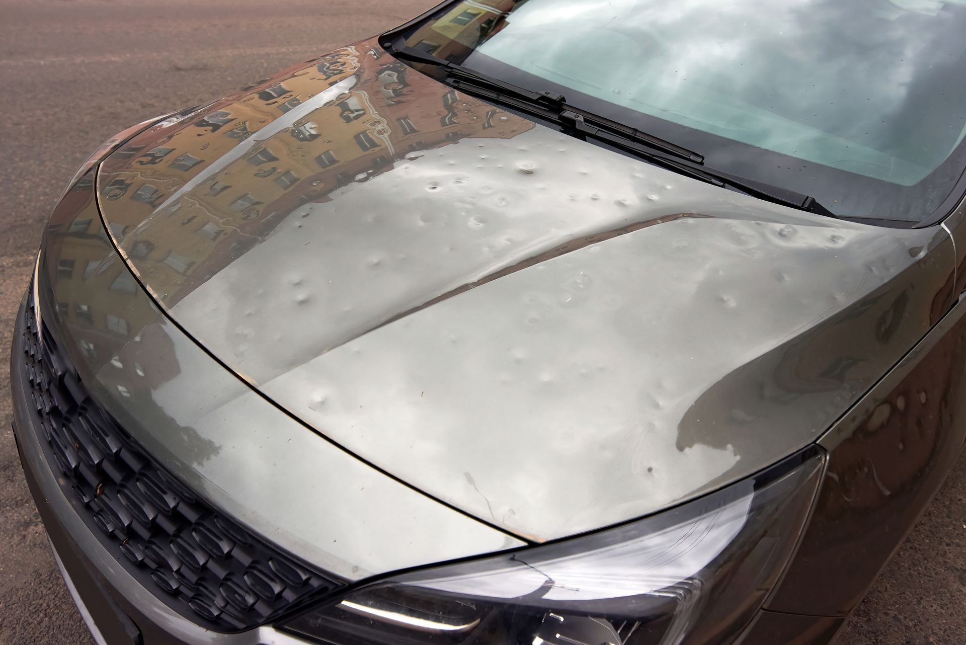 Silver car hood with visible hail dents and wet windshield parked outdoors