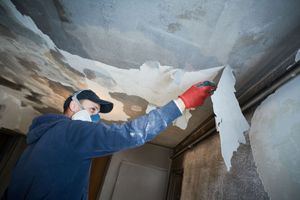 A person in a respirator mask and gloves peels old, damaged wallpaper from a ceiling.
