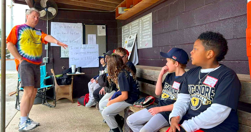A man in tie-dye shirt shares points with seated children wearing baseball t-shirts.