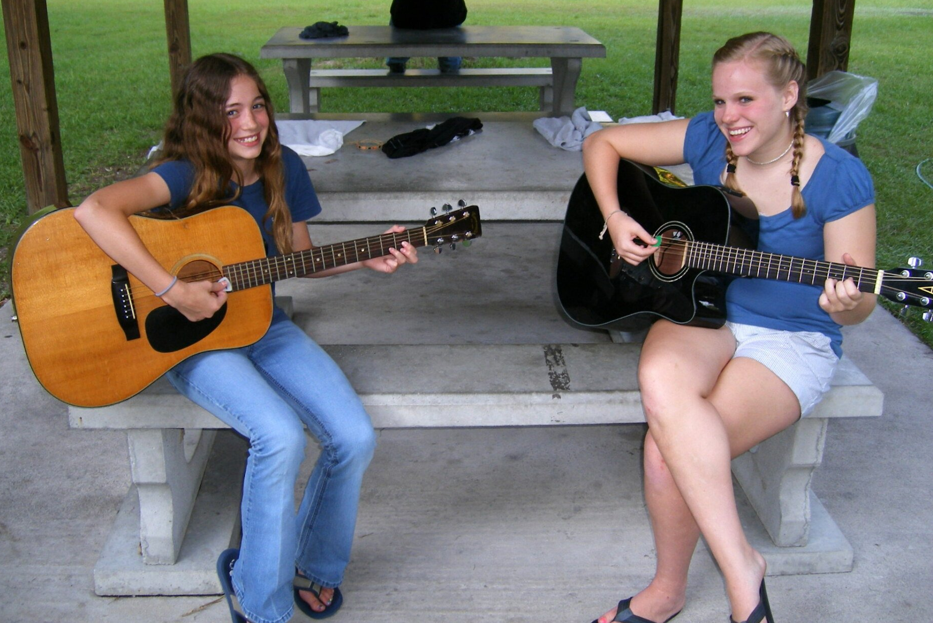 Two people playing acoustic guitars on a picnic table in a park, smiling casually