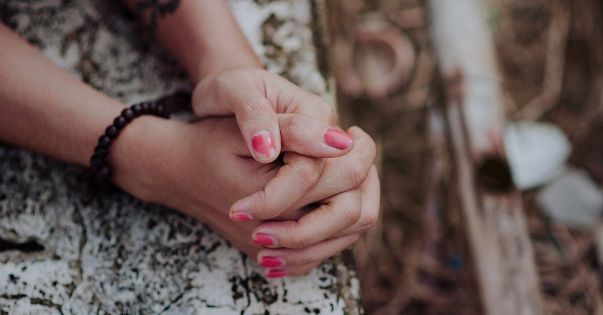 Hands clasped, wearing a beaded bracelet and pink nail polish, resting on patterned fabric.