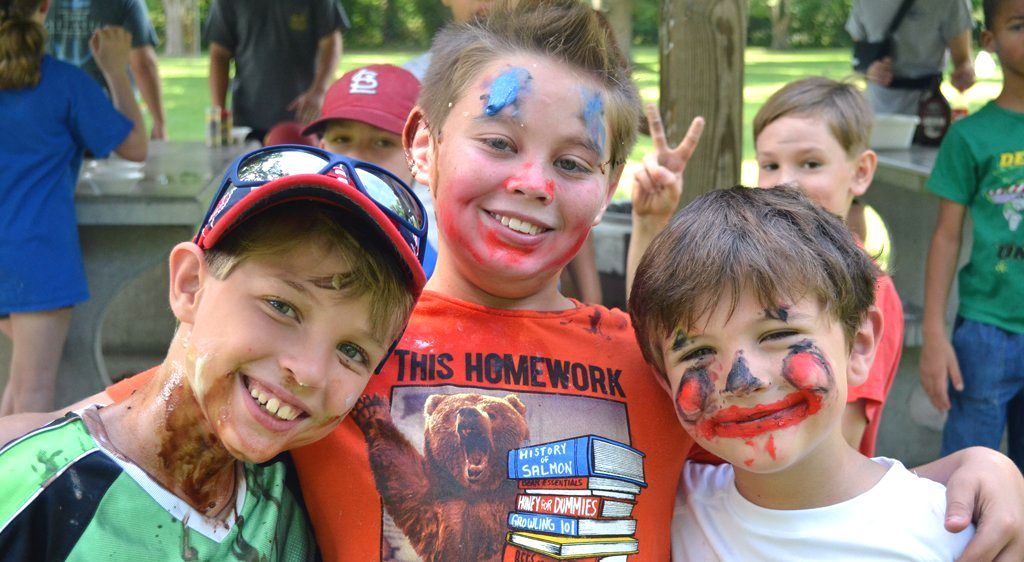 Smiling kids with face paint posing outdoors, one holding a homework sign and others making peace signs