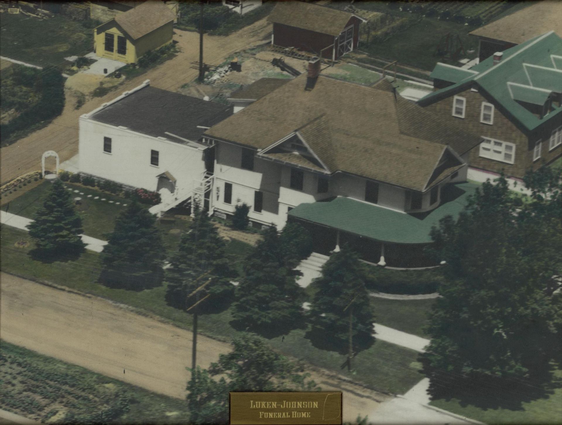 Aerial view of a white house with a brown roof and evergreen trees along a street.
