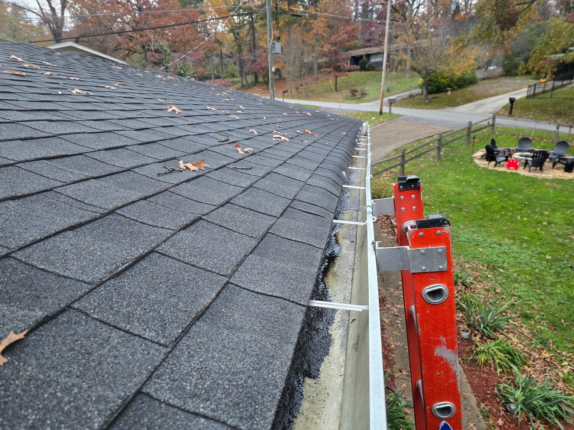 A view from a ladder looking along a shingled roof edge with a metal gutter, overlooking a lawn and neighborhood street.