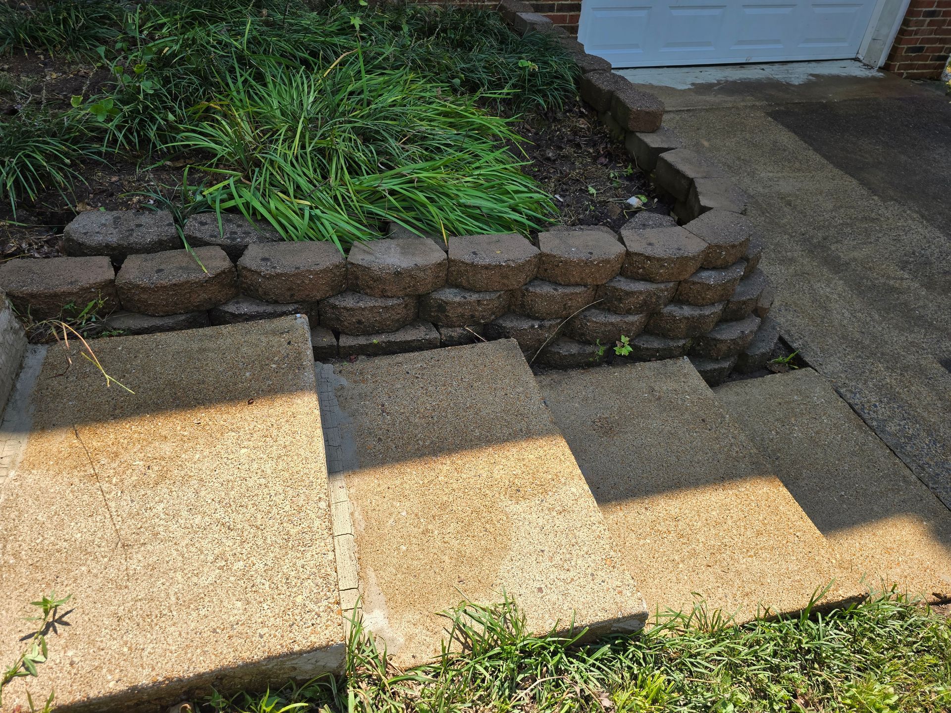 Three concrete steps lead up to a small garden bed enclosed by a stone retaining wall next to a paved driveway.