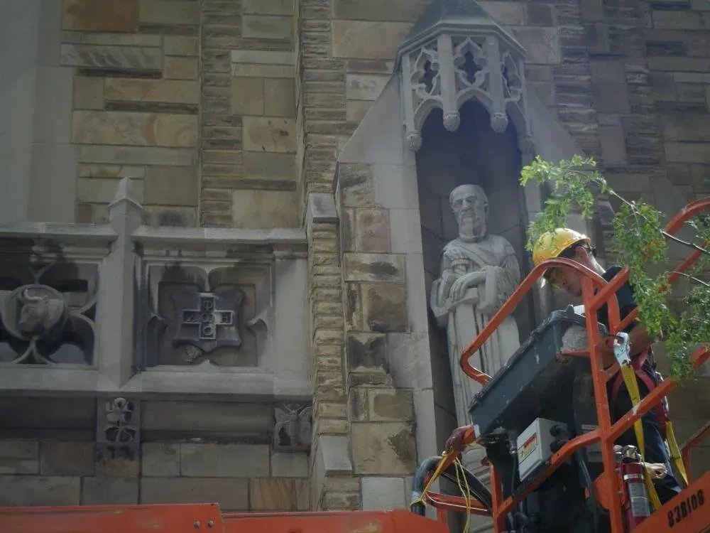 Worker on lift trims foliage near stone statue in gothic building niche.