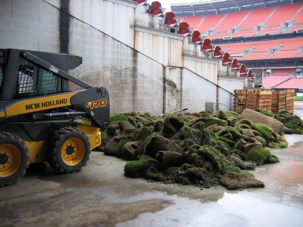 New Holland skid steer next to a pile of rolled-up sod near a stadium wall.