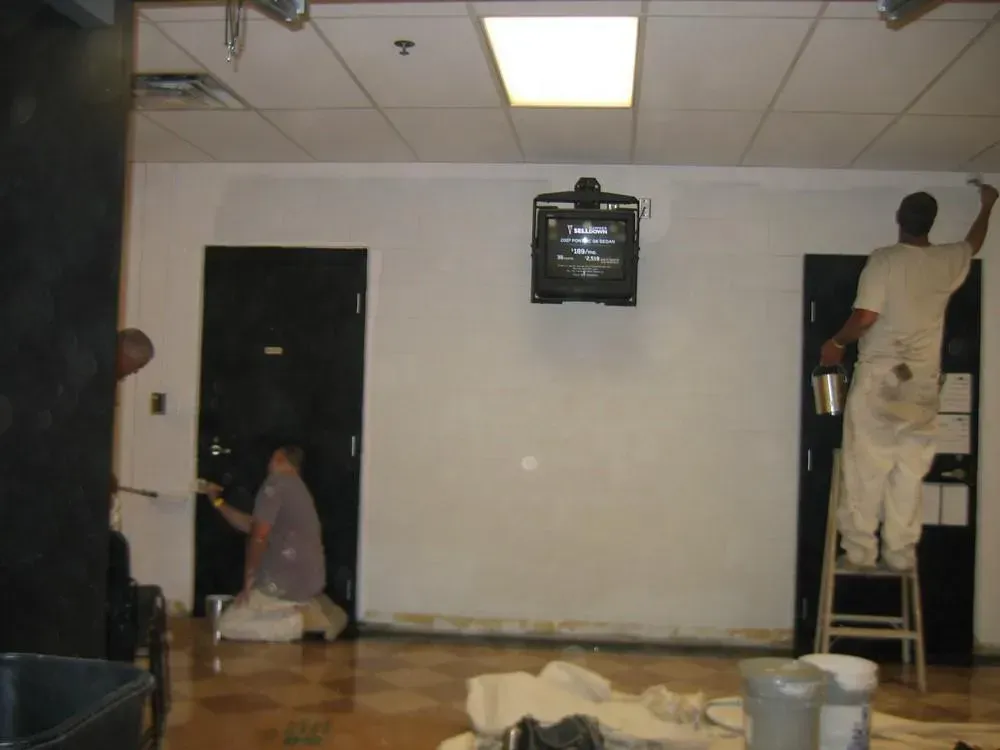 Two people painting a white wall in a room. One paints on a stool, the other crouches near a black door.