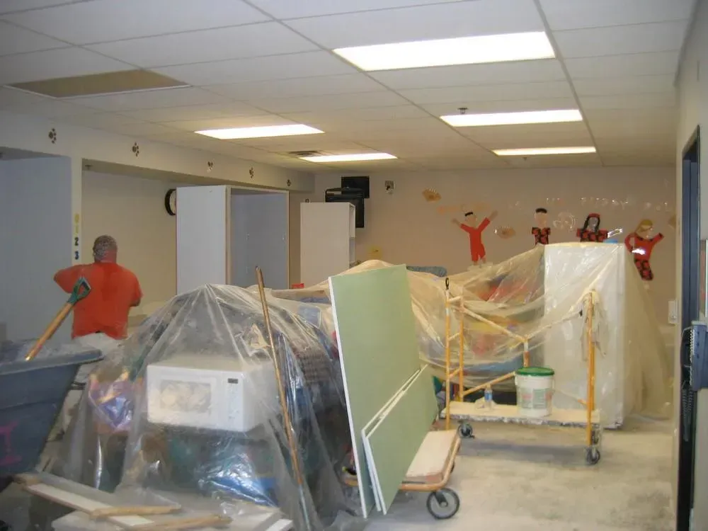 Construction site; man sweeping floor, covered appliances, drywall on cart, children's art on wall.