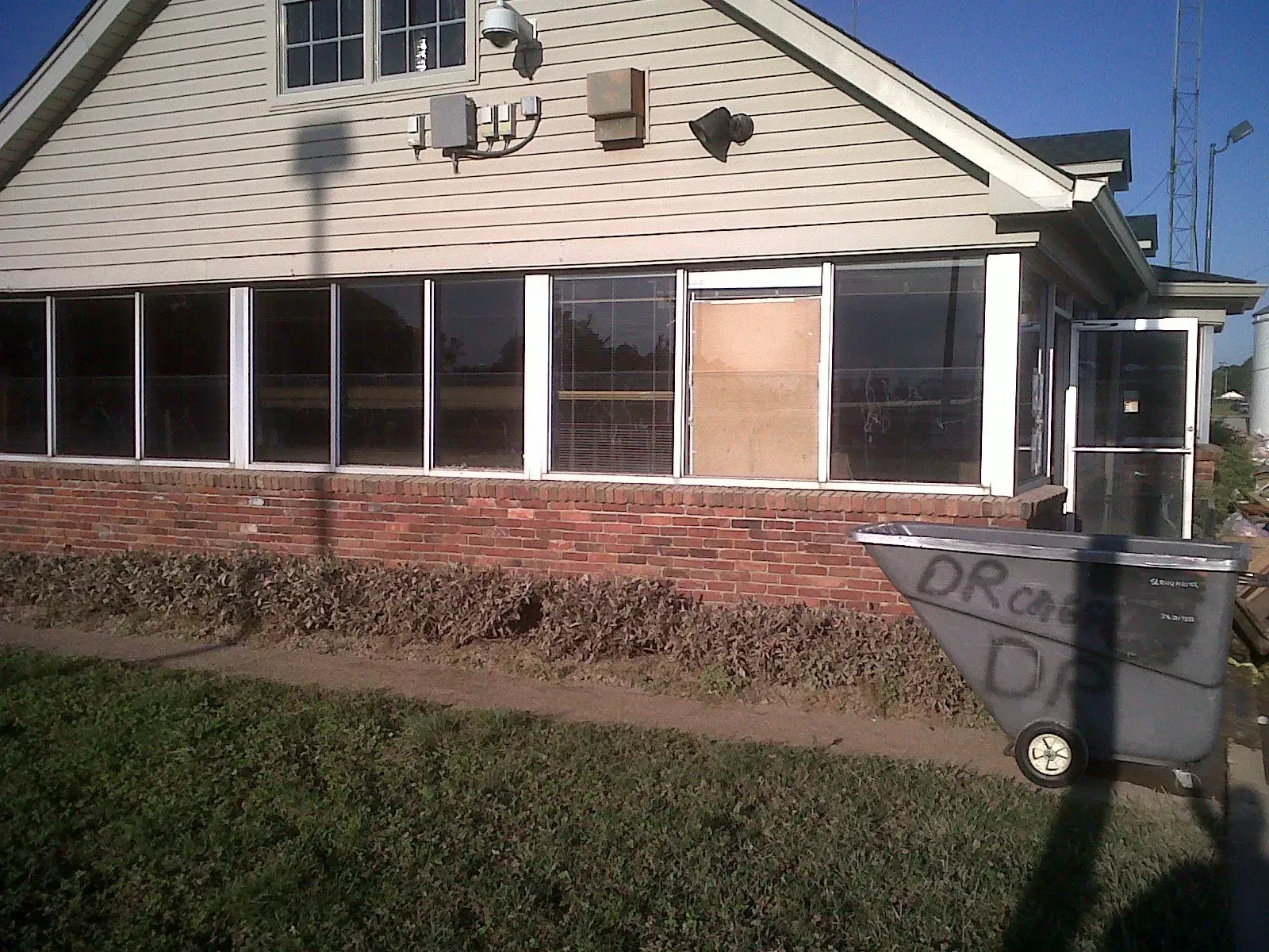 A light-colored building with a screened porch and brick base, a trash can in the foreground.
