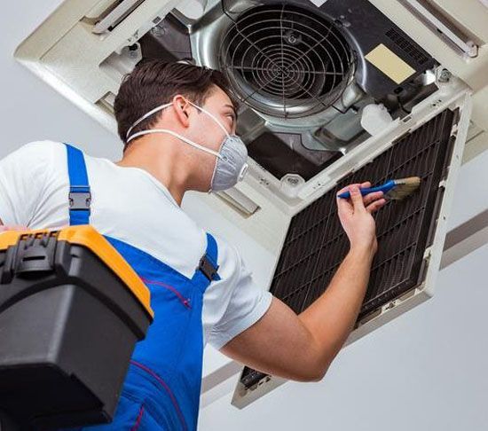 Man cleaning air conditioning unit filter with a brush, wearing mask and overalls.