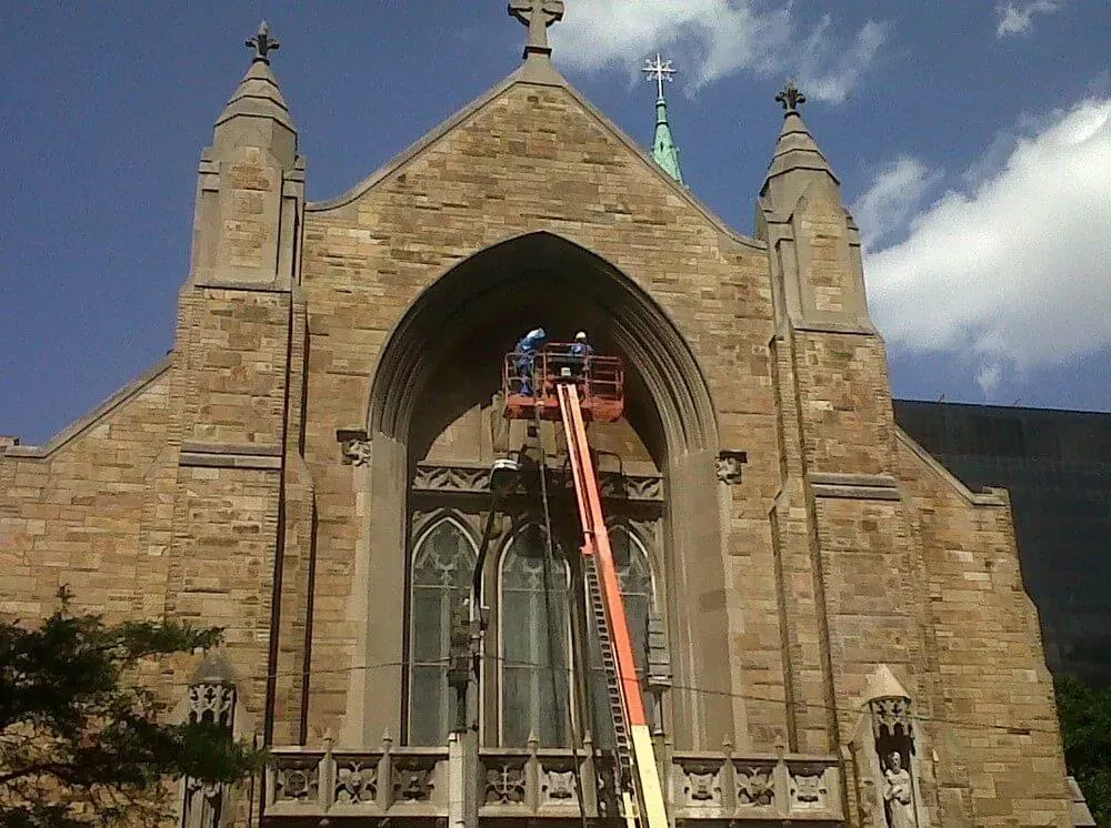 Church building with workers on a lift, repairing the arched window, against a blue sky.