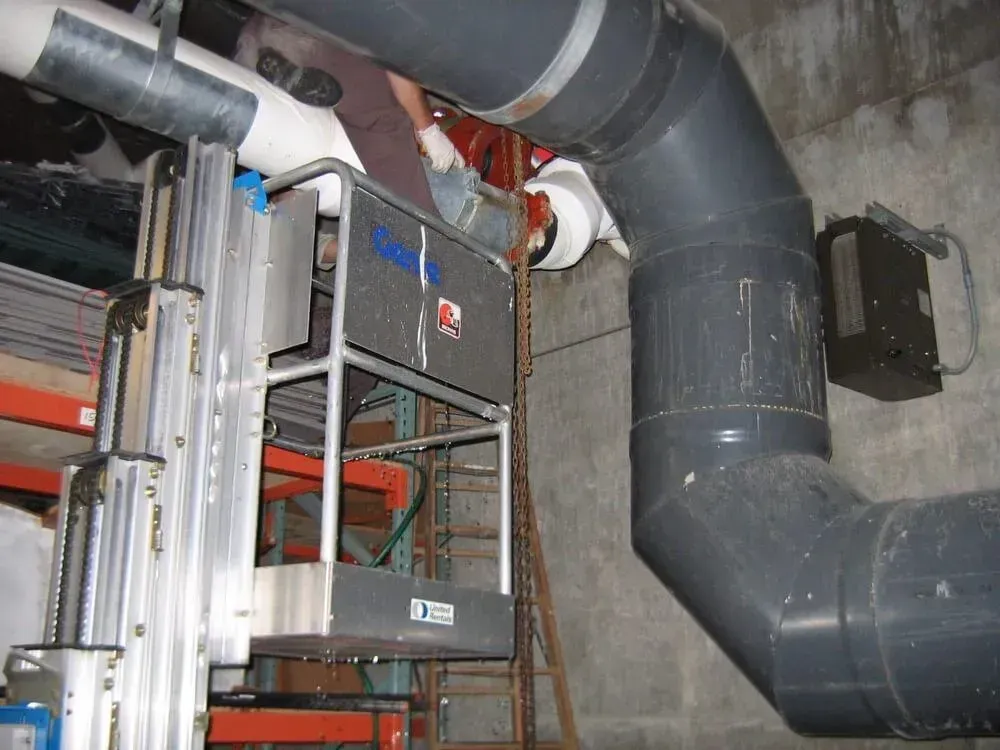 Worker in lift servicing large pipes in a concrete room. Gray and white pipes, storage shelving, and mounted equipment are visible.