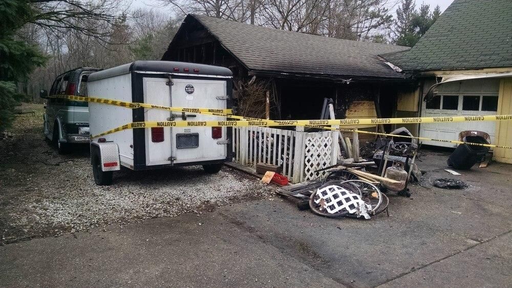 A house with fire damage is cordoned off with caution tape; a trailer and debris are nearby.