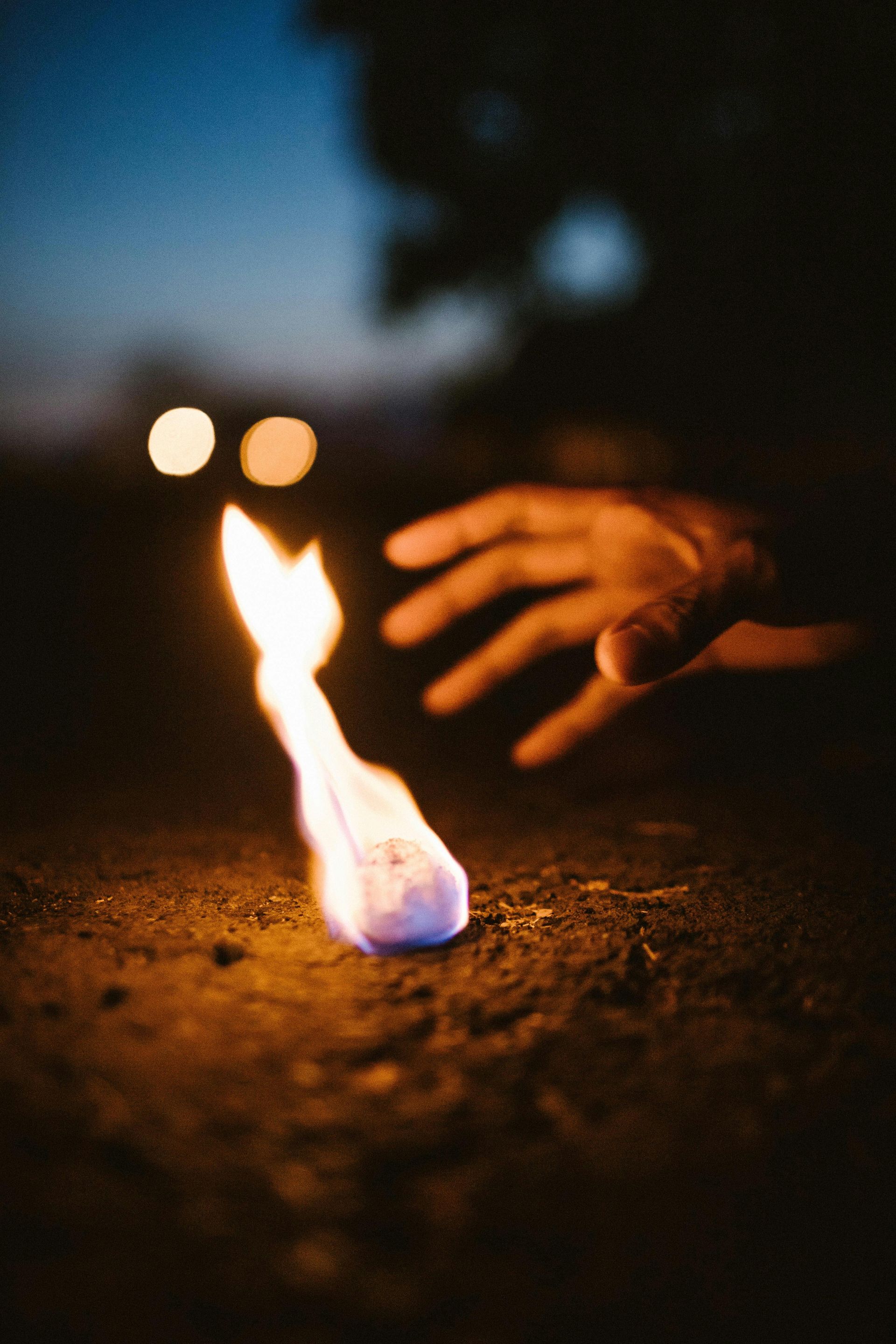 A hand reaching toward a flame burning on the ground at night.
