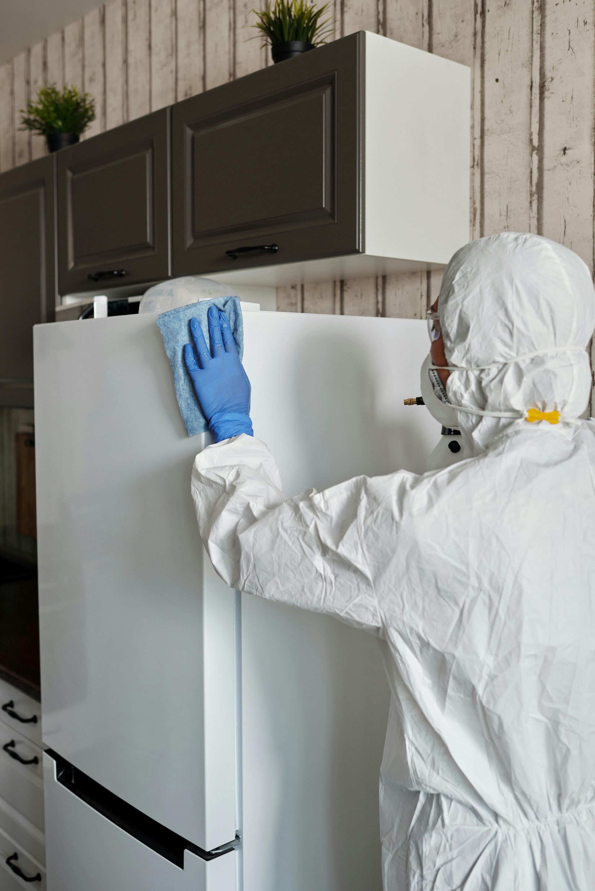 Person in protective suit cleaning a white refrigerator with a blue cloth in a kitchen.
