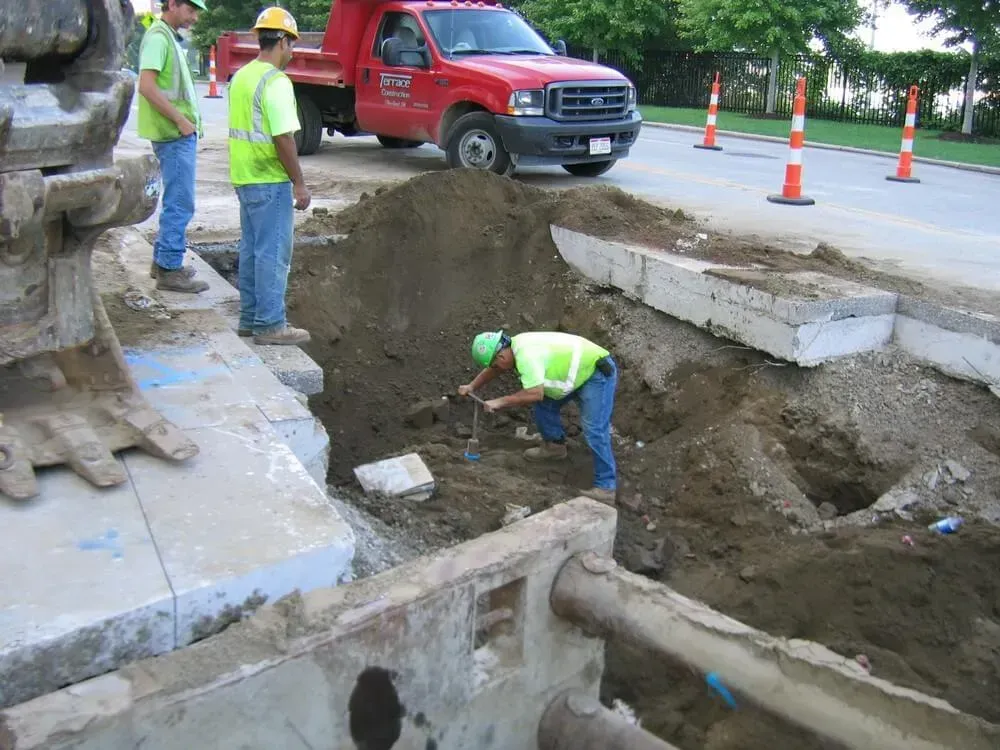 Construction workers excavating dirt, truck, concrete barriers, road cones.