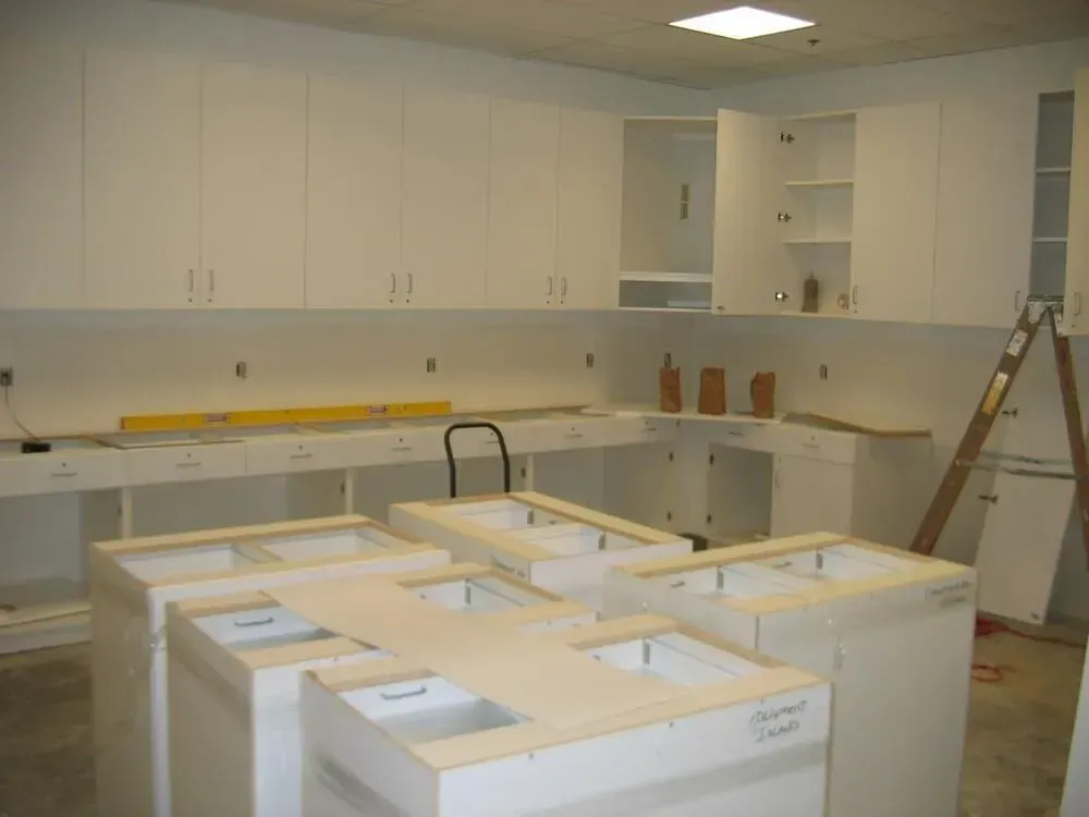 Kitchen cabinets being installed, with base cabinets in foreground and upper cabinets on wall. White cabinets.