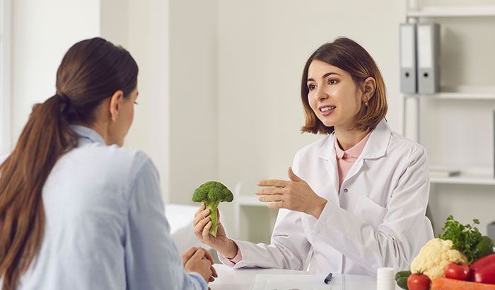 A woman is sitting at a table talking to a nutritionist while holding a piece of broccoli.