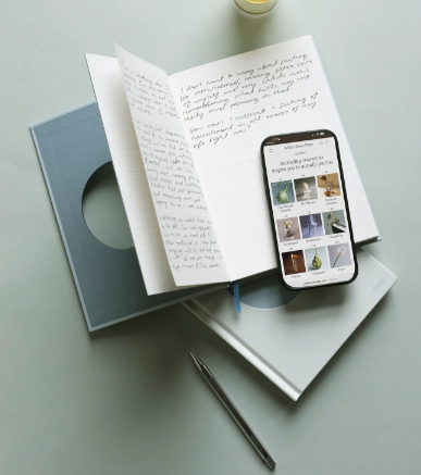 Open notebook with handwritten notes beside a smartphone displaying a photo gallery, on a pale desk