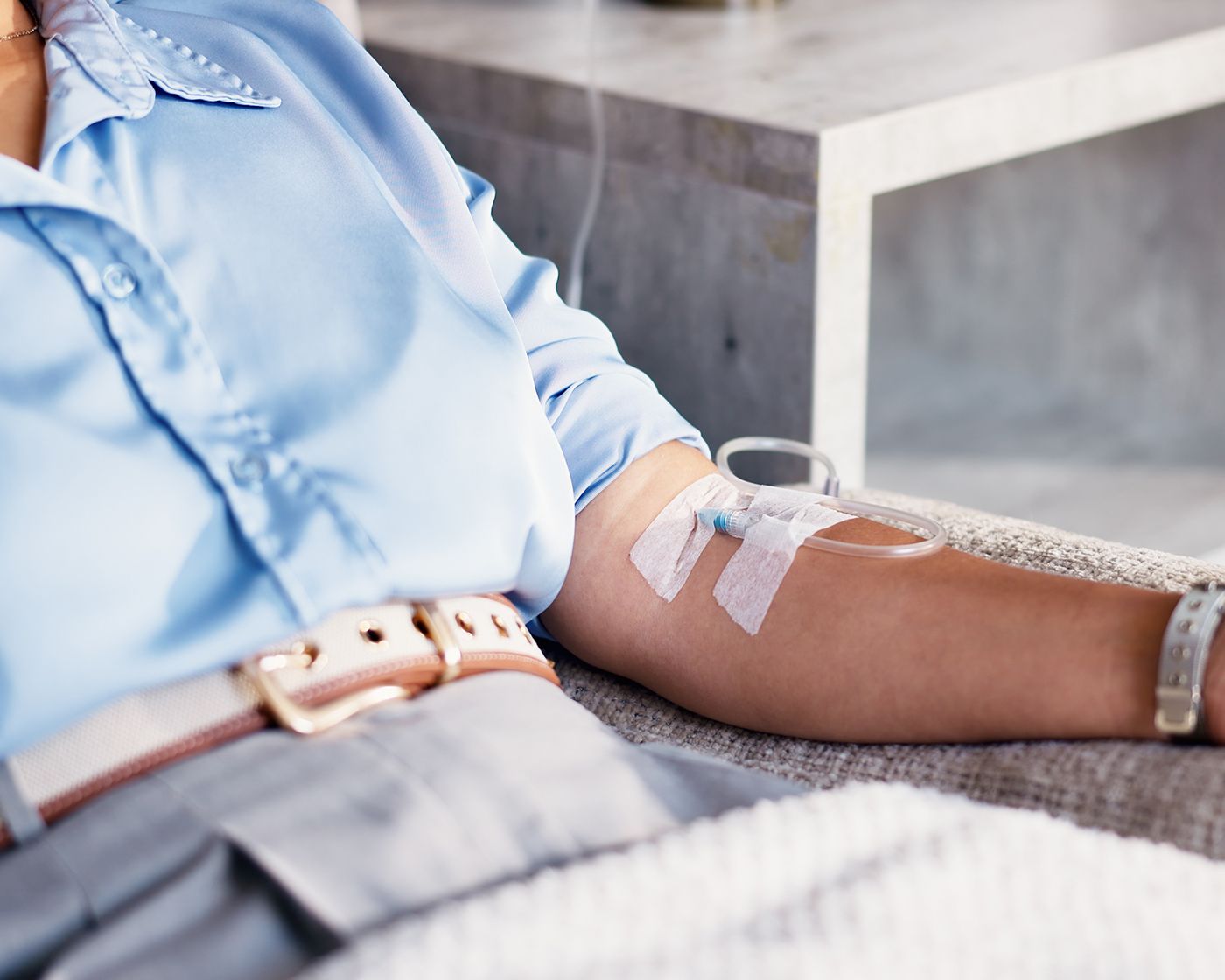 Arm with IV catheter and tape, resting on a couch, wearing a light blue shirt and gray pants