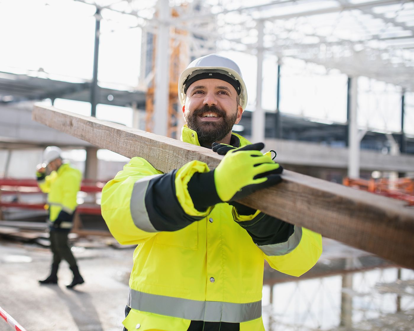 Construction worker in neon yellow safety gear carrying a wooden beam at an outdoor worksite.