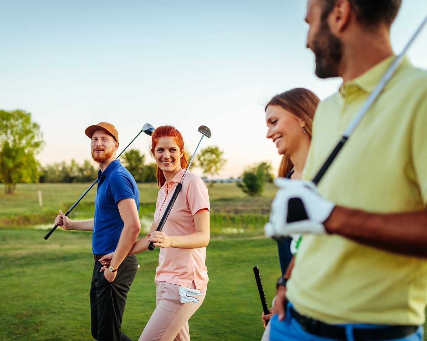 Four golfers walking on a green course, carrying clubs and chatting under a blue sky