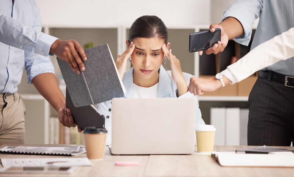 A woman is sitting at a desk in front of a laptop computer surrounded by people.