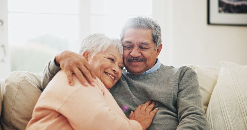 An elderly couple is sitting on a couch hugging each other.