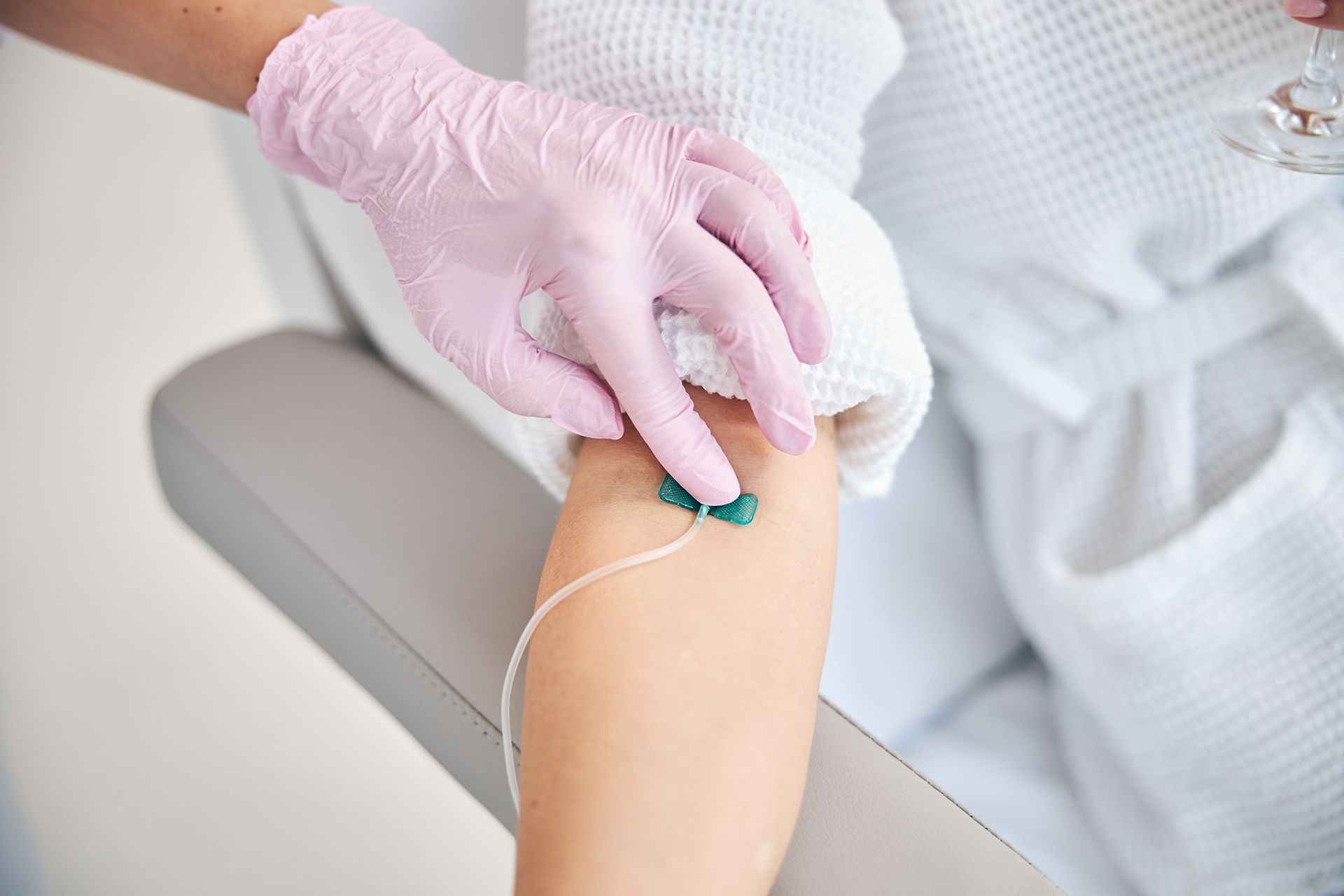 A gloved hand securing an IV line on a person's forearm, who is wearing a white bathrobe.