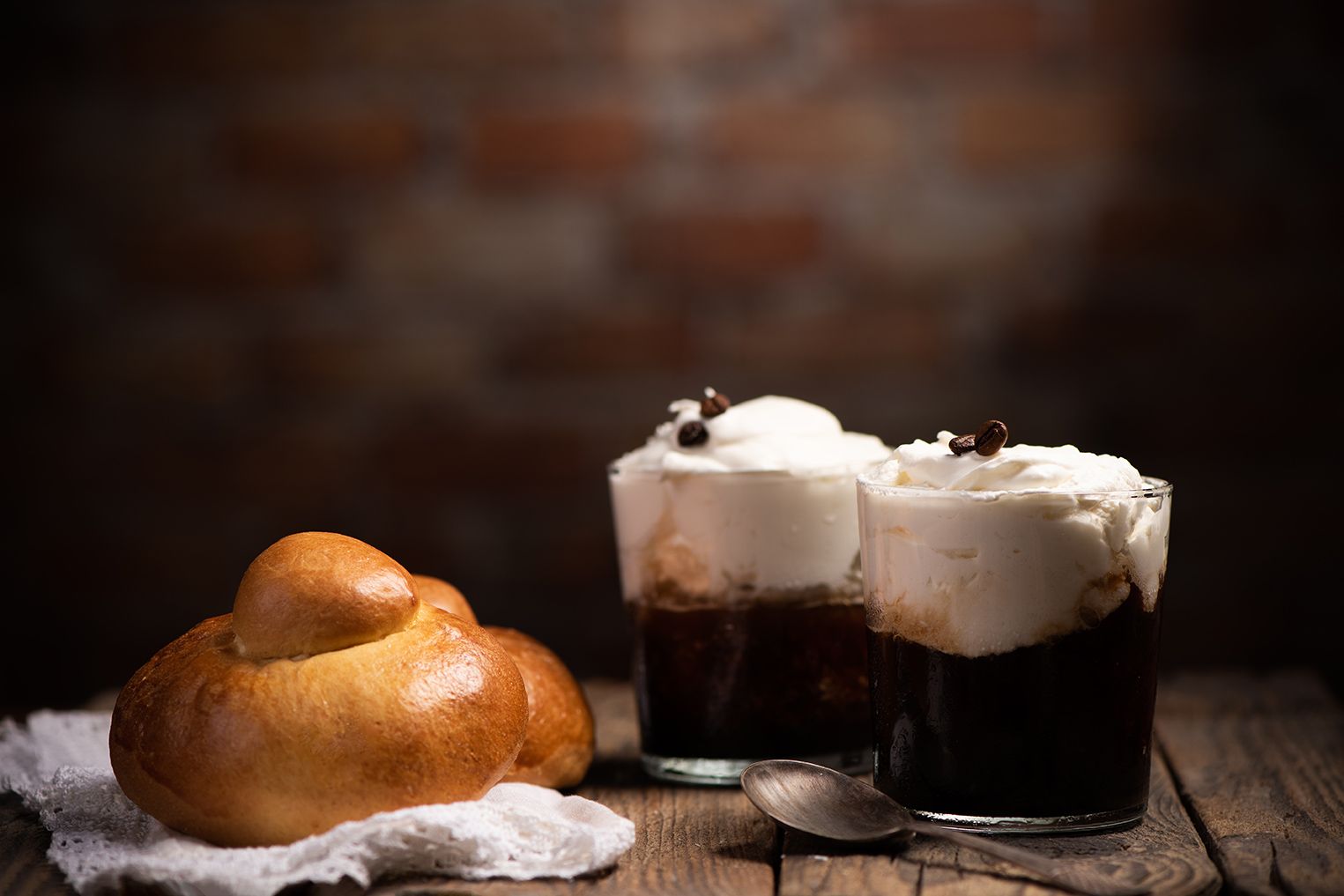 Panino brioche su un panno accanto a due bevande al caffè con panna montata, su un tavolo di legno, sfondo di mattoni.