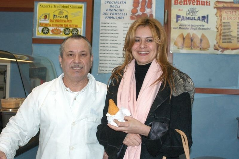 Un uomo in camice bianco e una donna sorridono. Sono accanto a un espositore di cibo. La donna tiene in mano un cono di pasta.