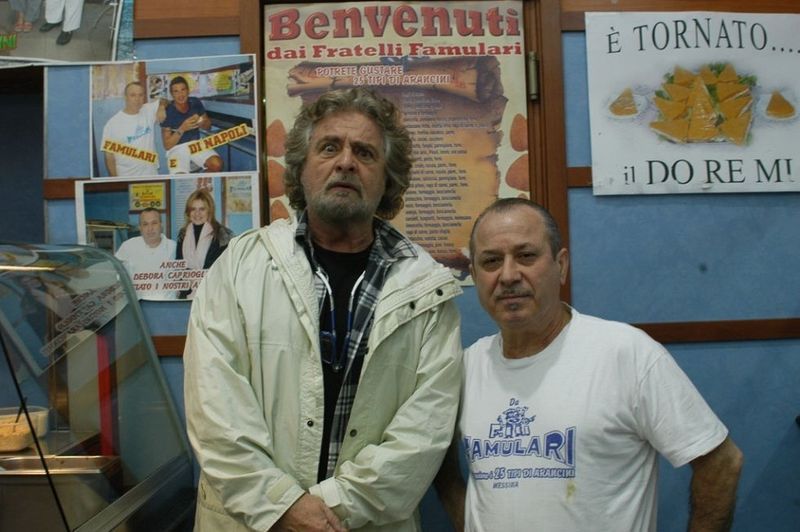 Due uomini posano in un negozio, uno indossa una giacca, l'altro una camicia bianca. Sullo sfondo l'insegna del negozio e alcune foto.