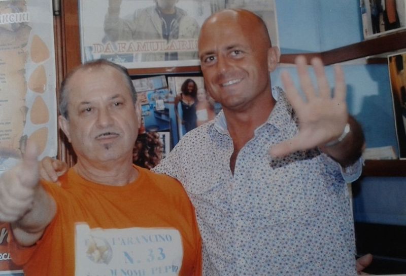 Due uomini sorridono, uno saluta con la mano. L'uomo a sinistra con la maglietta arancione fa il gesto del pollice in su. L'uomo a destra ha la testa calva e indossa una maglietta a fantasia.