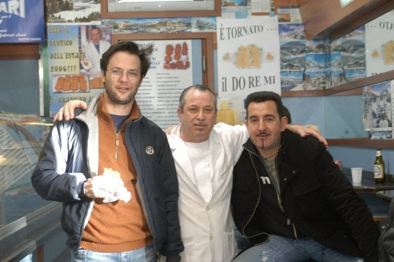 Tre uomini in posa; uno in camice bianco, affiancato da altri due. Interno del negozio, alcuni manifesti visibili.