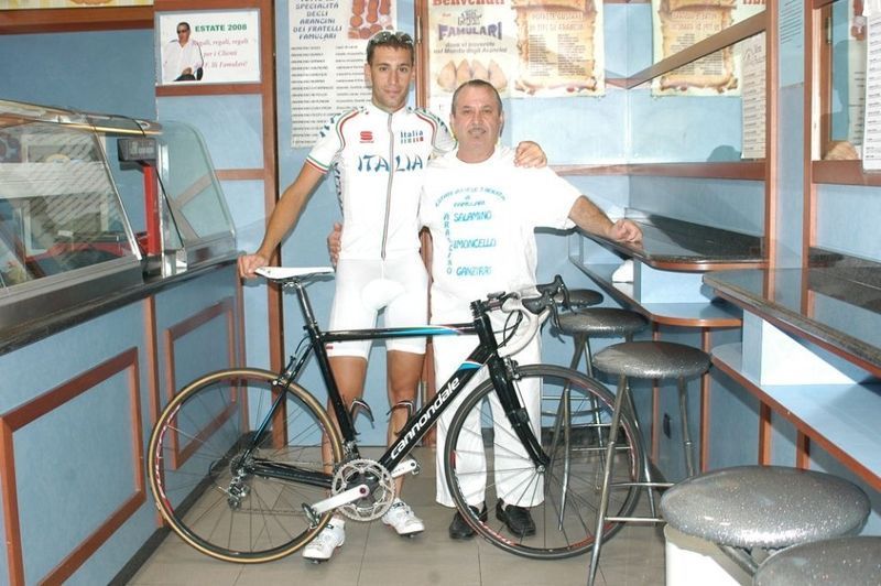 Ciclista con la maglia dell'Italia e uomo in posa accanto a una bicicletta Cannondale in un bar, pareti blu.