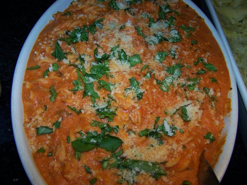 Piatto di pasta in una pirofila ovale bianca, ricoperto di salsa all'arancia, guarnito con erbe verdi e formaggio grattugiato.