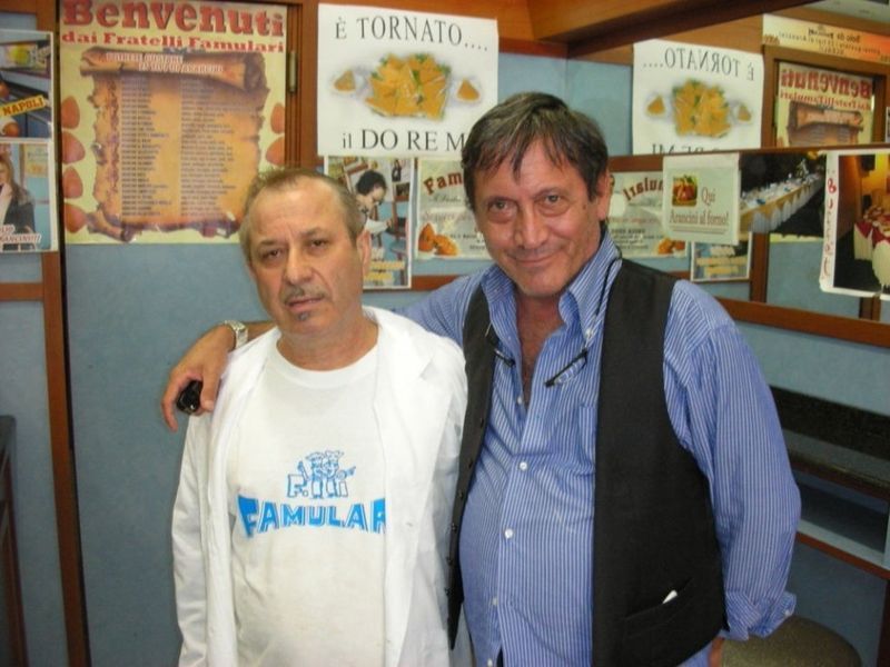 Due uomini in posa, uno con un braccio intorno all'altro. All'interno di un bar con poster di cibo e scritte in italiano.