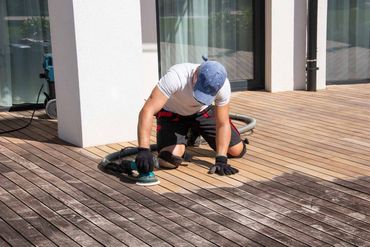 Person kneeling on a wooden deck, using a power tool near a white building.