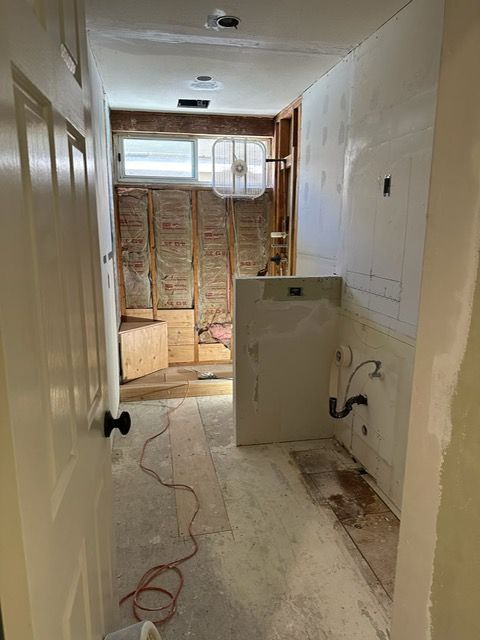 Narrow hallway under renovation, with exposed insulation, unfinished walls, and construction debris on the floor.