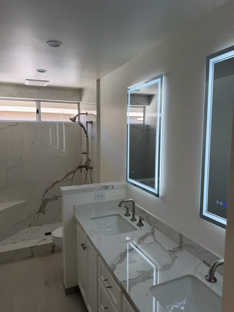 Modern bathroom with double sinks, lit mirrors, and a tiled shower with marble-pattern walls.