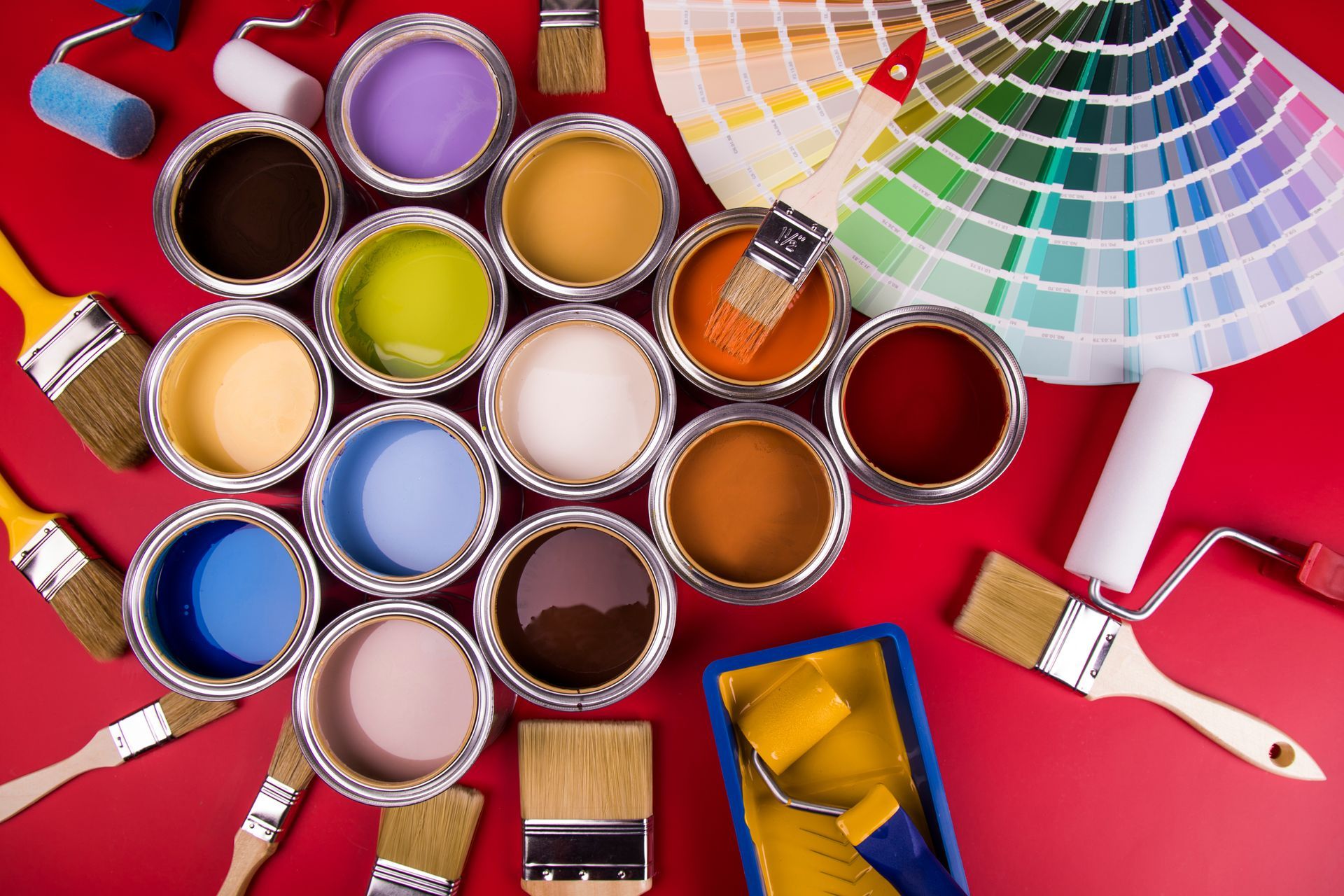Open paint cans with colors arranged on a red table beside brushes, rollers, and color swatches Open paint cans with colors arranged on a red table beside brushes, rollers, and color swatches