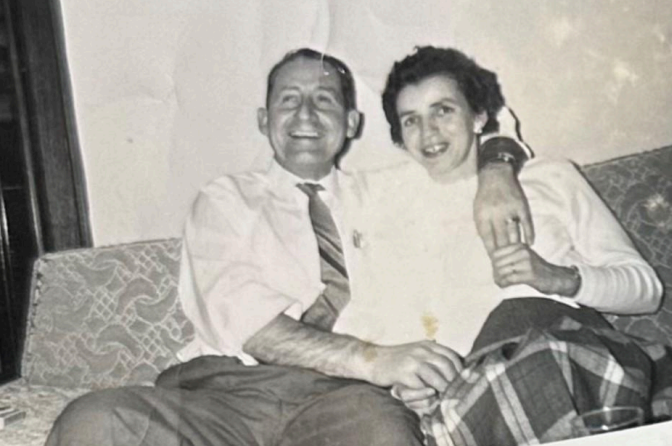 A couple sitting on a couch, smiling and embracing while looking toward the camera in a vintage black-and-white photo.