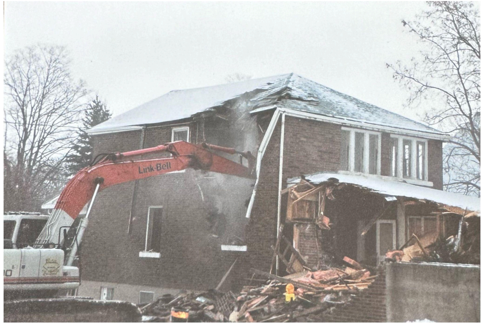 An orange excavator demolishing a brick house on a cloudy, wintry day.