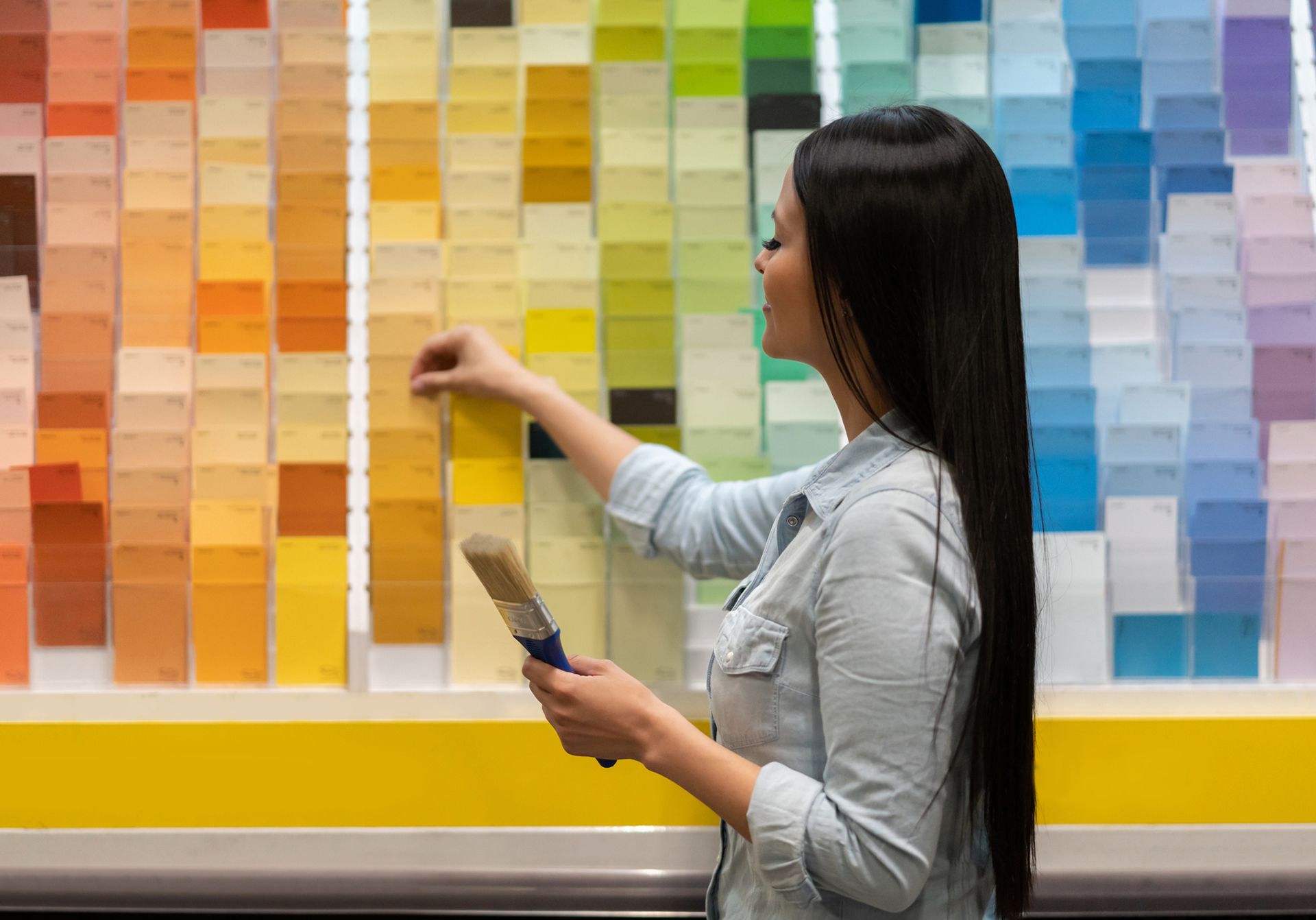 Woman choosing paint color from a display of swatches, holding a paintbrush. Woman choosing paint color from a display of swatches, holding a paintbrush.