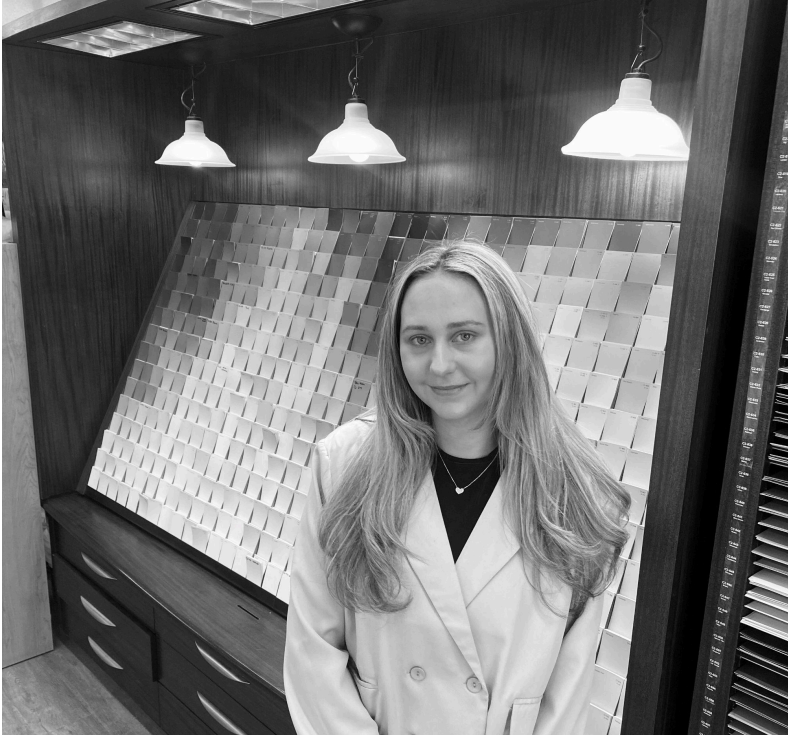 A person with long hair wearing a blazer stands in front of a wall display of small square paint samples under lights.