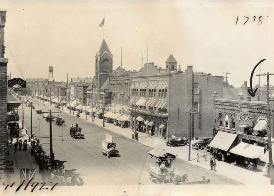 A vintage street scene in a town, featuring commercial buildings with awnings, pedestrians, and early motor vehicles.