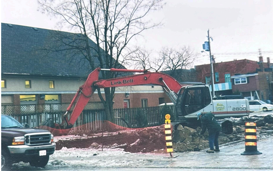 A red excavator parked in a snowy town lot near a brick building, with a worker standing nearby and orange safety cones.
