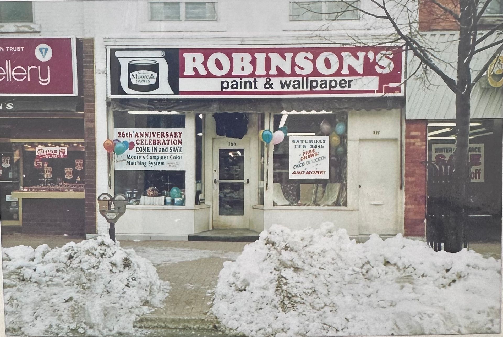 Storefront of Robinson's Paint & Wallpaper decorated with balloons for an anniversary, with snow piles in the foreground.